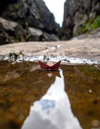 A tiny red paper boat embarks on a grand voyage across a small puddle, its reflection mirroring the vast sky above. Set against a rugged mountain canyon, this image captures a moment of quiet adventure, symbolizing hope, imagination, and the start of a new journey, no matter how small.の素材