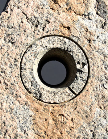 A perfectly circular hole is precisely drilled into the rugged, speckled surface of a granite slab. This macro view highlights the powerful intersection of industrial technology and raw, natural stone, creating a compelling abstract background of texture and form.の素材