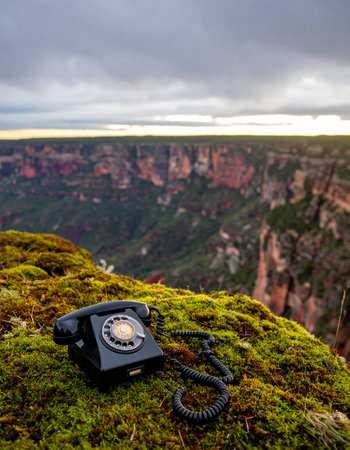 An old rotary phone rests on a moss-covered cliff, a forgotten relic overlooking a vast and silent canyon. It represents a surreal juxtaposition of technology and nature, evoking themes of lost connections, unanswered calls, and the profound silence of the wilderness.の素材