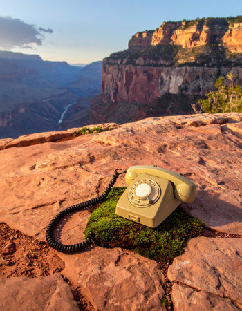 An old rotary telephone rests on a patch of moss at the edge of a vast canyon, a surreal and anachronistic sight. This conceptual image represents a lost connection, a call to nature, or the strange juxtaposition of old technology in a timeless, wild landscape, evoking feelings of nostalgia, mystery, and isolation.の素材