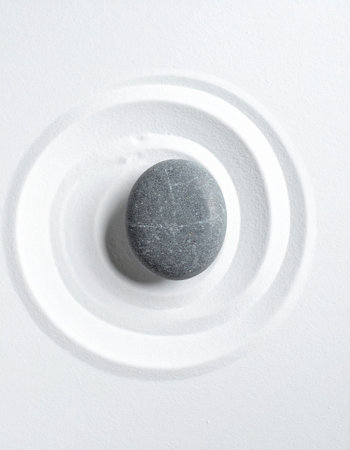 A single, smooth stone rests at the heart of a tranquil Zen garden. Concentric circles raked into the pure white sand emanate outwards, creating a visual metaphor for focus, mindfulness, and the peaceful ripple effect of a single point of calm. This minimalist composition evokes a sense of balance, harmony, and serene meditation.の素材