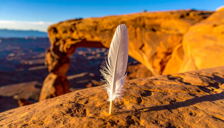 A single, delicate white feather stands in stark contrast to the rugged red sandstone, catching the first golden rays of sunrise. In the background, the iconic Mesa Arch frames a vast desert landscape, evoking a sense of peace, freedom, and the gentle touch of nature in a powerful environment.の素材