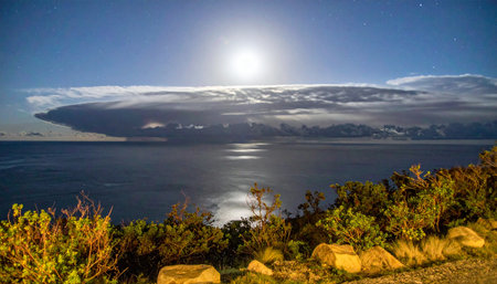 A brilliant full moon casts a shimmering path of light across the calm ocean surface. Above, a vast and dramatic cloud formation stretches across the night sky, creating a breathtaking and otherworldly celestial spectacle over the quiet, rocky shore.の素材