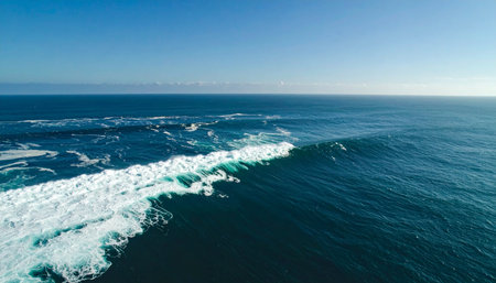 From high above, the immense power of the ocean is on full display as a massive wave curls and breaks, sending white foam across the deep blue sea. A scene of raw, untamed nature and majestic beauty under a clear sky.の素材