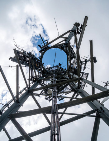 A low-angle view captures the intricate and chaotic framework of an industrial tower reaching into an overcast sky. At its heart, a large, circular blue dish reflects the heavens, serving as a focal point in this complex web of steel and technology, hinting at powerful communication systems or a relic of a futuristic past.の素材
