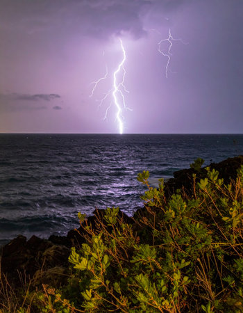 Under a violet, stormy sky, a brilliant lightning strike connects heaven and earth over the restless sea. This awe-inspiring display of natural energy captures a moment of intense power and dramatic beauty, viewed from the safety of a rocky shore.の素材