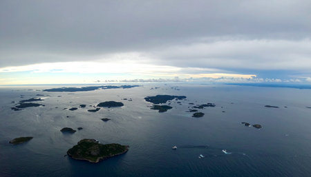 From a high vantage point, countless green islands dot the vast, tranquil ocean. A dramatic, overcast sky looms above, with a sliver of bright light on the horizon, suggesting a coming storm or the break of dawn over this remote and untouched natural paradise.の素材
