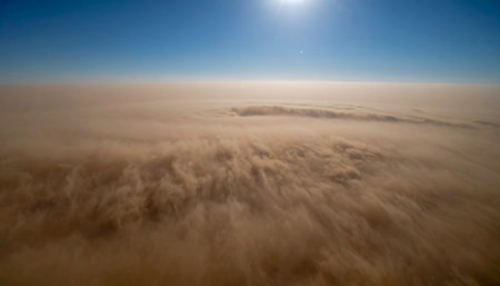 From a high vantage point, a massive and powerful dust storm churns across the landscape, a turbulent sea of sand under a clear blue sky. The sun shines brightly above, illuminating the immense scale of this dramatic and awe-inspiring natural weather event, symbolizing the raw force of nature.の素材