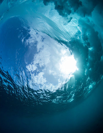 From beneath the surface, the world transforms. A powerful ocean wave curls overhead, creating a temporary cathedral of water and light. The sun bursts through the translucent blue, illuminating the turbulent beauty in a moment of serene power and natural wonder.の素材