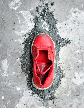 A single, vibrant red childs shoe rests on a pile of dark ash, a stark symbol of lost innocence and forgotten memories. The desaturated, gritty concrete background emphasizes a feeling of loneliness and abandonment, leaving the viewer to wonder about the story behind the missing pair.の素材