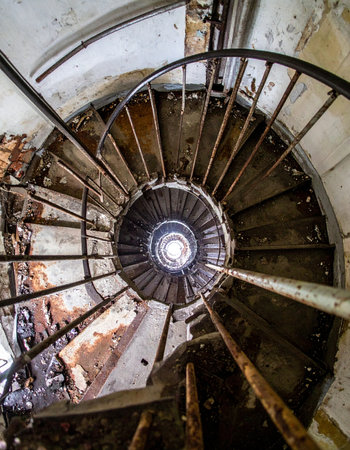 A low-angle view captures the haunting beauty of a decaying spiral staircase. Each rusty, worn step tells a story of forgotten times, leading the eye upward towards a distant circle of light, symbolizing hope, escape, or an unknown future in a derelict building.の素材