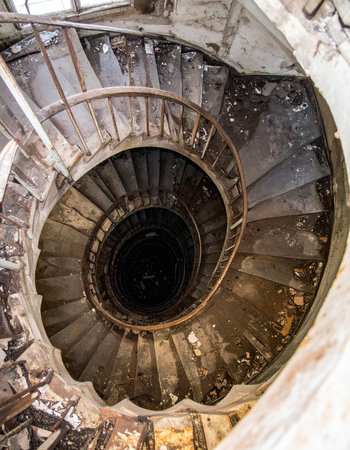 Looking down from the top of a forgotten tower, a decaying spiral staircase winds its way into a dark, unknown abyss. Each worn and rusted step tells a story of times relentless passage, creating a sense of mystery, suspense, and the eerie beauty of urban decay.の素材