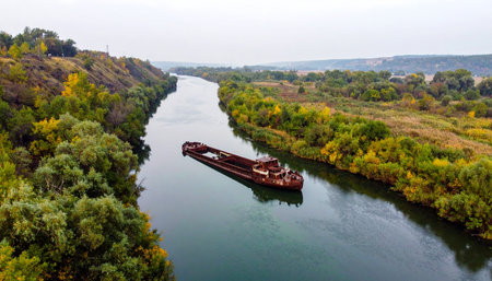 An aerial perspective captures a solitary cargo barge as it glides silently down a winding river. Flanked by dense forests transitioning into the warm colors of autumn, the scene highlights the vital role of inland waterways in commerce and the serene beauty of industrial transport meeting the natural world.の素材