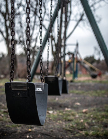 An empty swing set sits silently in a deserted park on a cold, overcast day. The scene evokes a powerful sense of nostalgia and melancholy, reflecting on forgotten childhood memories, the quiet passage of time, and the feeling of being left behind.の素材