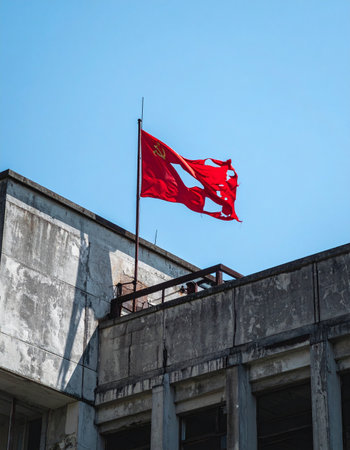 A tattered red flag, its fabric torn by wind and time, flies defiantly against a clear blue sky. Perched atop a weathered, stark building, it stands as a poignant symbol of past struggles, faded glory, and enduring resilience. The scene captures a moment of quiet history and the relentless passage of time.の素材