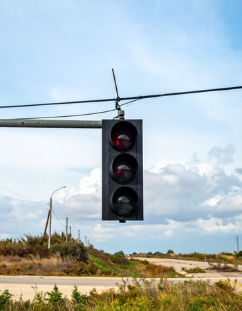 A lone traffic light hangs suspended over a quiet country road, its unlit signals a silent sentinel against a vast, cloud-filled sky. This image captures a moment of stillness and anticipation at an empty intersection, symbolizing transition, control, and the quiet infrastructure that governs our journeys.の素材