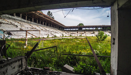 From the shadows of a ruined press box, a view of what once was a field of dreams. Now, nature has taken over, with wild grass and weeds covering the ground where athletes once competed. The silent, crumbling stands are a testament to a forgotten era, a powerful symbol of decay and the relentless passage of time.の素材