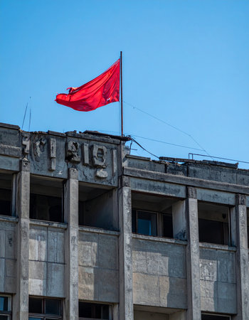 A vibrant red flag, a potent symbol of past ideologies, waves defiantly against a clear blue sky. It stands atop a weathered and decaying concrete building, its empty windows like hollow eyes staring into history. This powerful image evokes themes of resilience, memory, and the enduring legacy of political change.の素材