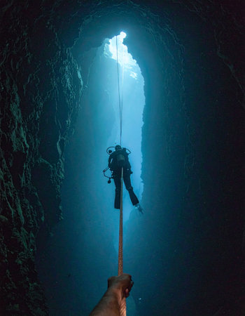A lone scuba diver ascends on a guide rope through the deep, tranquil blue waters of an underwater cave. A brilliant beam of sunlight pierces the darkness from an opening above, illuminating the path to the surface and symbolizing hope, discovery, and the end of a challenging journey into the unknown.の素材