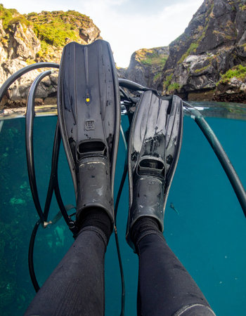 From a first-person perspective, a scuba divers fins rest at the tranquil surface of the water, splitting the view between the clear blue depths below and the rugged cliffs of a secluded cove above. A moment of calm anticipation before descending into the underwater world.の素材