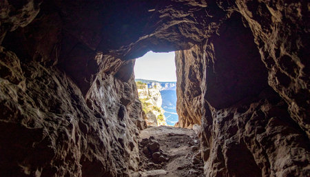 A dramatic perspective from within a dark, textured rock cave, looking out through a natural opening. The bright light at the end of the tunnel reveals a stunning coastal scene with a brilliant blue sea, suggesting themes of hope, discovery, and escape into natures beauty.の素材
