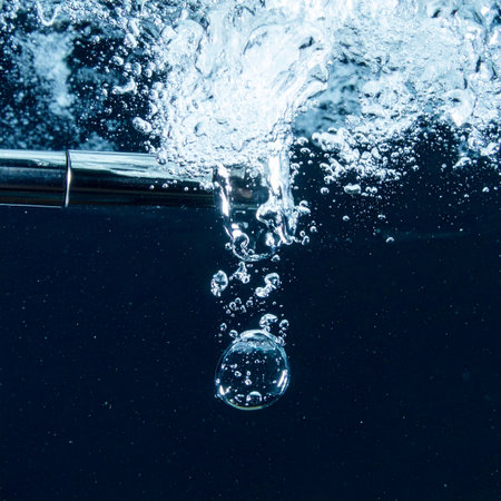 A powerful jet of clean water bursts from a submerged metal pipe, creating a dynamic splash and a cascade of effervescent bubbles. This image captures a moment of pure energy and refreshment, symbolizing purification, technological processes, and the vital force of water.の素材