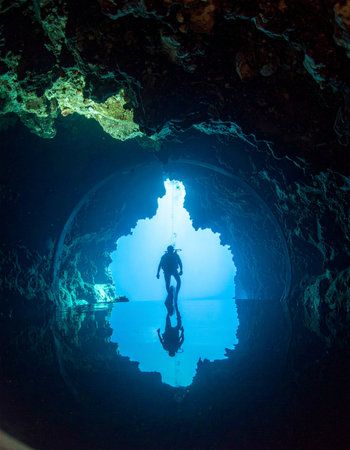 A lone scuba diver pauses at the glowing entrance of a majestic underwater cenote. The brilliant blue light illuminates the cavern, creating a stunning reflection and a sense of awe-inspiring adventure and serene discovery in the hidden depths of the earth.の素材
