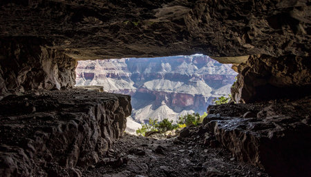 A breathtaking view of the vast Grand Canyon is perfectly framed by the dark, textured walls of a natural rock cave. This unique perspective offers a sense of discovery and solitude, looking out from a hidden shelter into the immense, sunlit landscape, highlighting the contrast between shadow and light.の素材