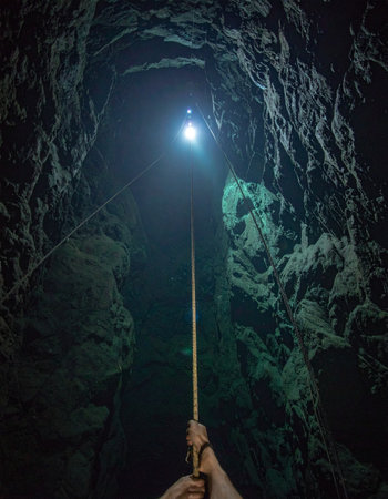From a first-person perspective, an explorer holds a long pole with a bright light, piercing the deep darkness of a narrow underground cavern. The beam illuminates the rough, textured rock walls, revealing the path forward into the unknown. This image captures the thrilling suspense and spirit of adventure in discovering hidden, subterranean worlds.の素材