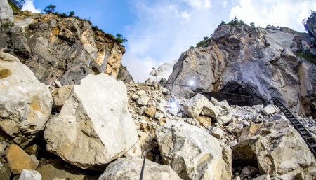 A low-angle view captures the immense scale of a mountain quarry, where massive boulders and stone debris tumble down the rugged cliff face. Sunlight illuminates the scene, highlighting the raw textures of the rock and the industrial nature of mineral extraction.の素材