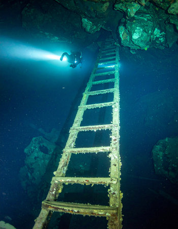A lone scuba diver illuminates a mysterious, submerged ladder deep within an underwater cave. The powerful beam of light cuts through the darkness, revealing a surreal scene that hints at a forgotten path and the thrill of exploring the unknown depths.の素材