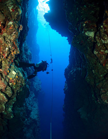 A lone scuba diver descends into the mesmerizing blue abyss of a narrow underwater canyon. Sunlight filters from the surface, illuminating the path into the mysterious depths, creating a sense of awe, adventure, and solitary exploration of the unknown.の素材