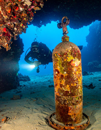 A scuba diver with a bright torch explores the mysterious depths of a sea cave, illuminating a long-forgotten, coral-encrusted air tank. This scene captures a moment of discovery, adventure, and the passage of time beneath the waves.の素材
