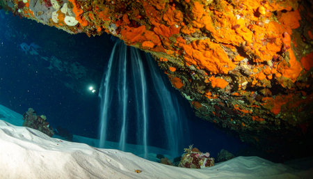 A lone diver discovers a breathtaking, surreal sight deep within a submerged cavern. A cascade of fine white sand flows like a waterfall from the rock ceiling, a rare geological wonder hidden in the silent, dark blue depths, evoking a sense of mystery and adventure.の素材