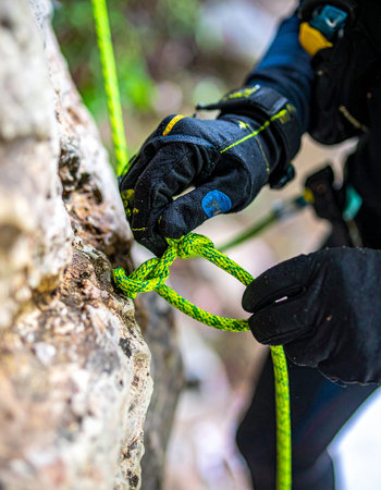 With focused precision, a climbers gloved hands expertly tie a critical safety knot into a vibrant green rope against a rugged rock face. This moment of careful preparation captures the essence of trust, skill, and the quiet concentration required before embarking on a vertical adventure.の素材