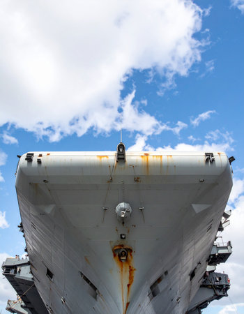 From a dramatic low angle, the immense bow of a historic aircraft carrier looms against a bright blue sky with scattered clouds. The weathered steel and streaks of rust tell stories of countless voyages and service, symbolizing national strength, naval power, and enduring history.の素材