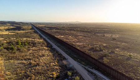 An aerial perspective captures the immense scale of the border wall as it cuts a stark line across the rugged desert terrain. Bathed in the warm glow of a rising sun, the structure stretches endlessly toward the horizon, symbolizing division, security, and complex international issues.の素材