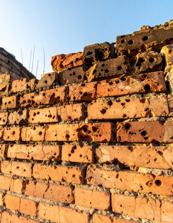A low-angle view captures the stark reality of a bullet-scarred brick wall, its weathered surface telling a story of past conflict and destruction. Against the backdrop of a clear, peaceful blue sky, the crumbling structure stands as a silent monument to resilience and the enduring passage of time.の素材