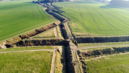 From a high angle, a network of historic military trenches carves deep lines into the peaceful green landscape. This powerful aerial perspective reveals the lasting scars of conflict and the ongoing work of archaeological excavation, uncovering stories of the past hidden just beneath the surface.の素材