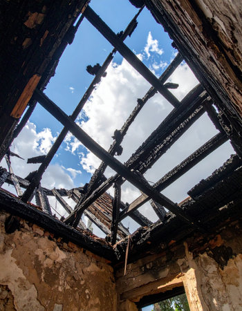 Looking up from the ashes of a devastating fire, the charred and skeletal remains of the roof frame a view of the bright blue sky. A poignant scene of destruction and loss, yet the open sky suggests resilience, hope, and the possibility of rebuilding from the ruins.の素材