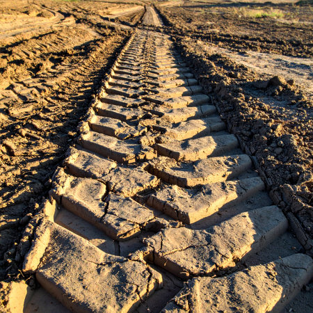 A close-up, low-angle perspective of a heavy vehicles tire track deeply imprinted into dry, cracked earth. The path leads into the distance, evoking concepts of industry, construction, progress, and the rugged journey forward.の素材