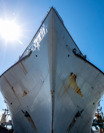 From a dramatic low-angle perspective, the immense bow of a naval vessel towers against a brilliant blue sky. The sun flares brightly, highlighting the ships powerful and weathered form, suggesting a history of long voyages and enduring strength.の素材
