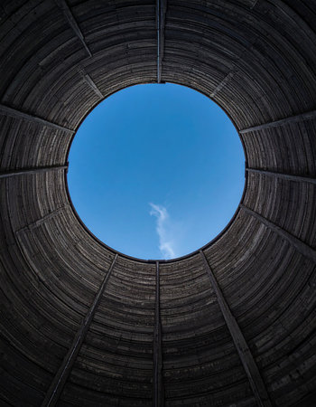A dramatic low-angle view from inside a dark, textured circular structure, looking up towards a perfect circle of bright blue sky. This powerful image symbolizes hope, freedom, and the concept of finding light at the end of the tunnel, representing opportunity and a breakthrough.の素材
