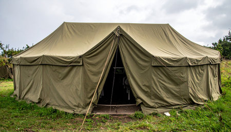 A sturdy, vintage canvas tent stands ready in a grassy field under an overcast sky. This rugged shelter evokes a sense of history, adventure, and preparedness, suitable for concepts ranging from military encampments and historical reenactments to outdoor survival and humanitarian aid.の素材