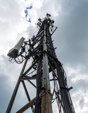 From a low angle, a complex cellular network tower stands tall against a dramatic, cloudy sky. Its intricate web of antennas, dishes, and cables represents the backbone of modern global communication, silently transmitting data and connecting people across vast distances.の素材