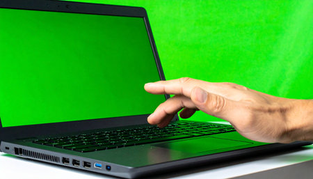 A person's hand reaches out to interact with a modern laptop, its screen glowing with a vibrant green chroma key.の素材