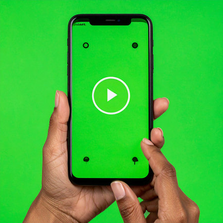 A close-up view of a person's hands holding a smartphone. The thumb is poised to tap the play button on the green chroma key screen, which is ready for custom content.の素材