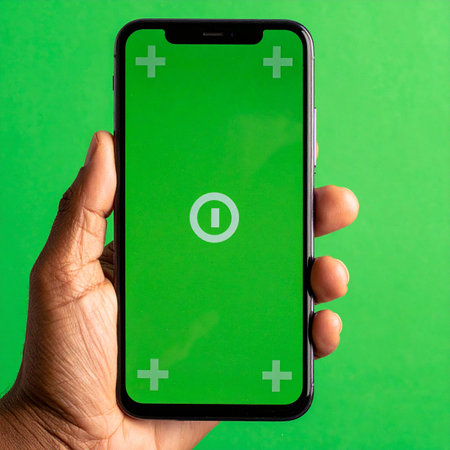 A close-up of a person's hand holding a modern smartphone, presenting its green screen with tracking markers.の素材