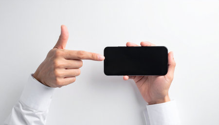 A top-down view of a person in a white shirt holding a smartphone horizontally and pointing to its blank screen.の素材