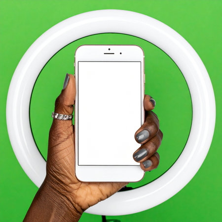 A content creator's hand holds up a smartphone with a blank screen, perfectly framed by a ring light for optimal lighting.の素材